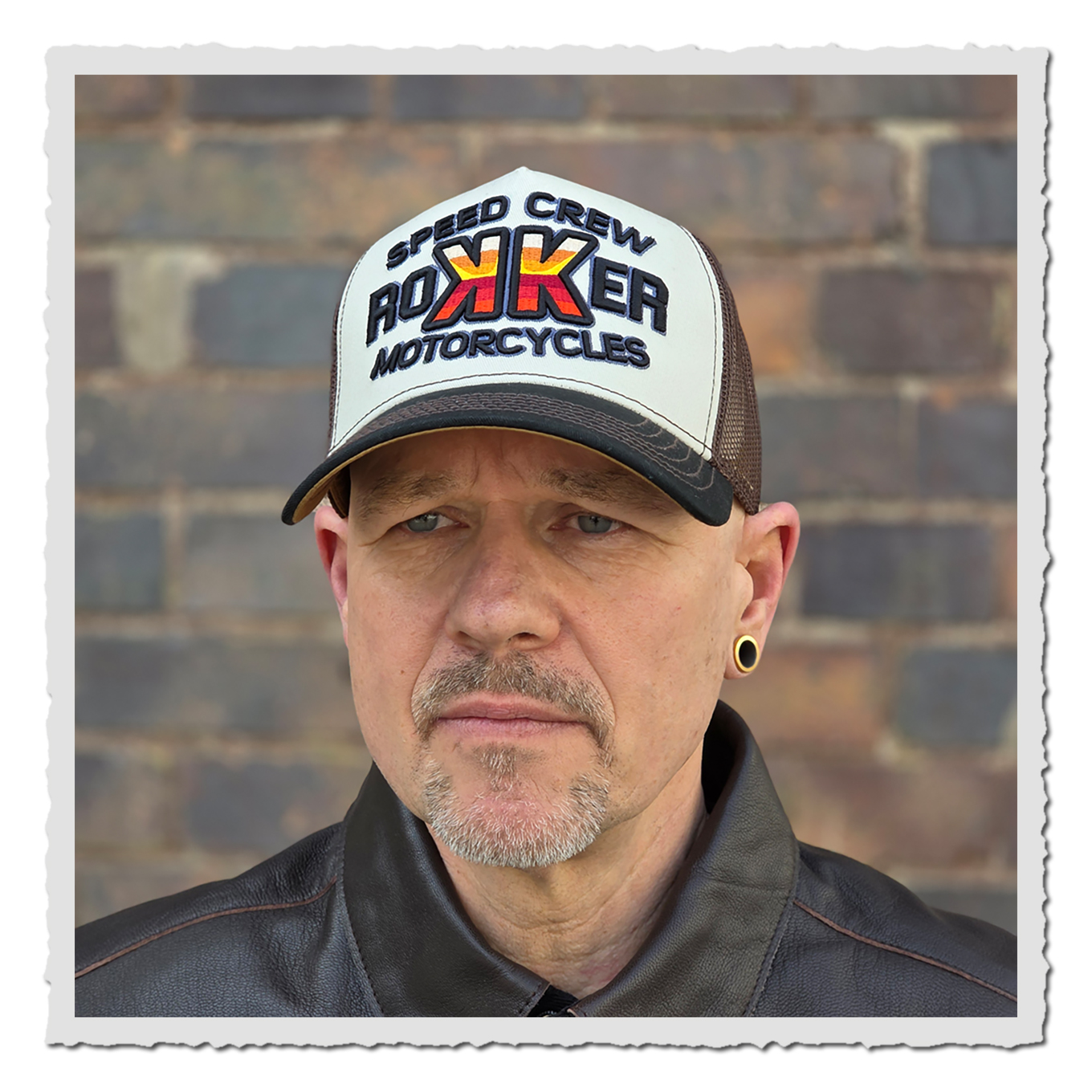 Nahaufnahme einer Person mit heller und brauner Cap vor Backsteinwand, Gesicht und Jackenkragen sichtbar.